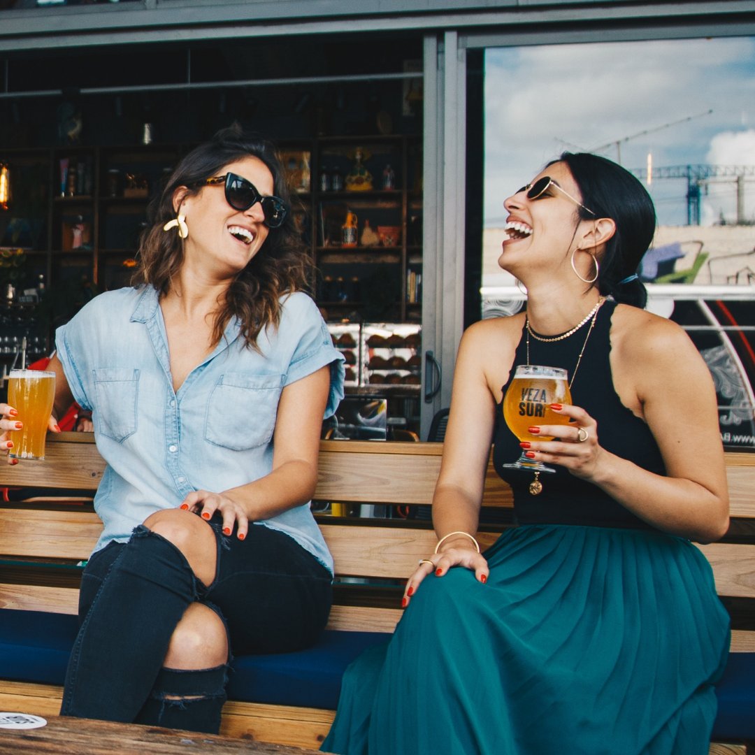 Duas mulheres sentadas num banco e dando gargalhadas. Elas seguram copos de cerveja.