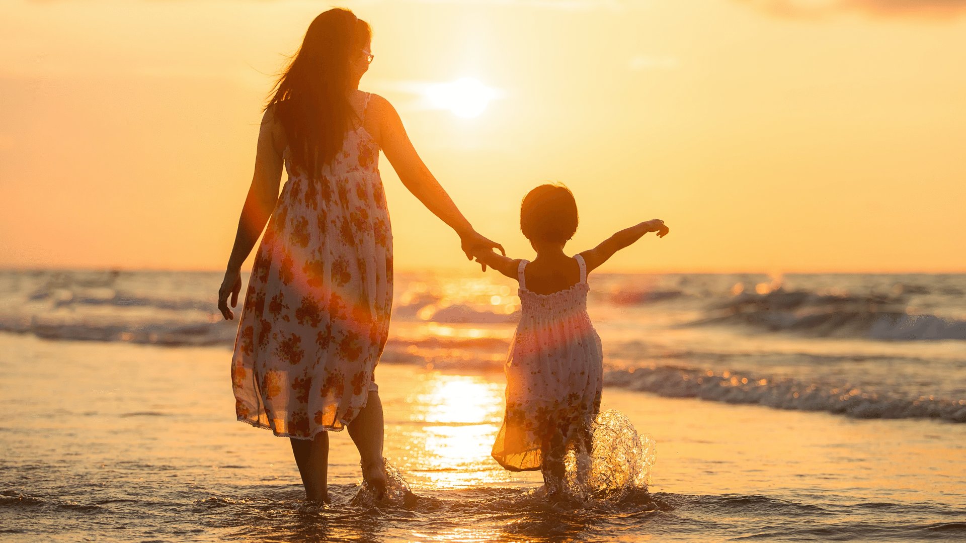 Mãe com a mão dada para sua filha criança enquanto as duas entram no mar com o sol poente ao fundo