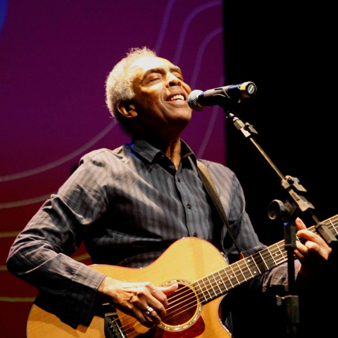 Imagem do cantor e compositor Gilberto Gil tocando violão e cantando em um palco