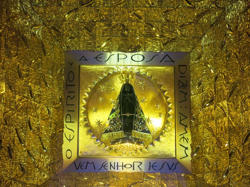 A imagem de Nossa Senhora Aparecida guardada em um receptáculo de vidro anexado em uma parede dourada, é possível ler as palavras 'A esposa', 'Dizem: amém', 'Vem Senhor Jesus' e 'O espírito'.