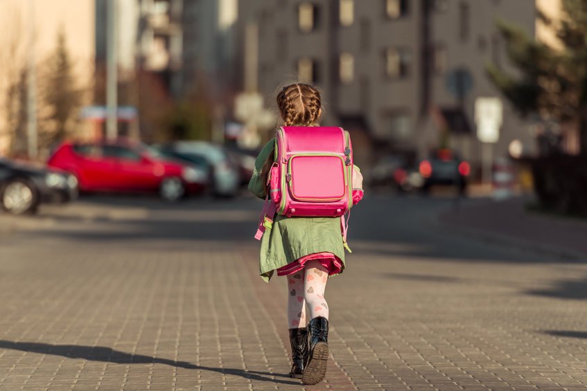 Criança andando na direção contrária da câmera usando vestido, meia-calça, botas e uma mochila.