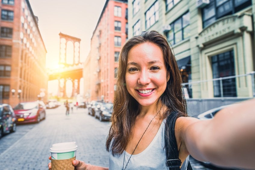 Mulher na rua tirando selfie segurando um café enquanto o sol nasce