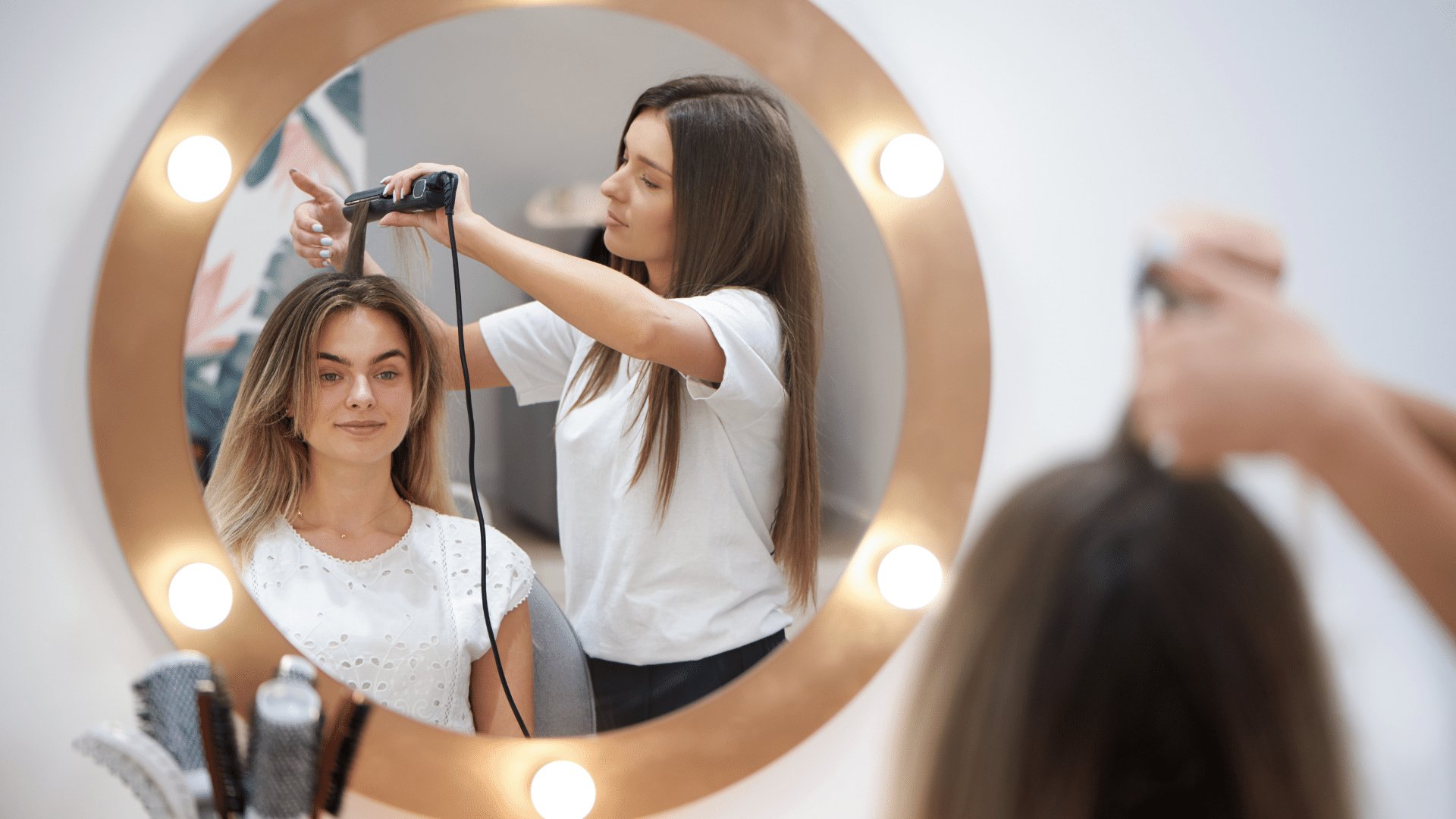 Espelho mostrando reflexo de uma mulher passando chapinha no cabelo de outra moça