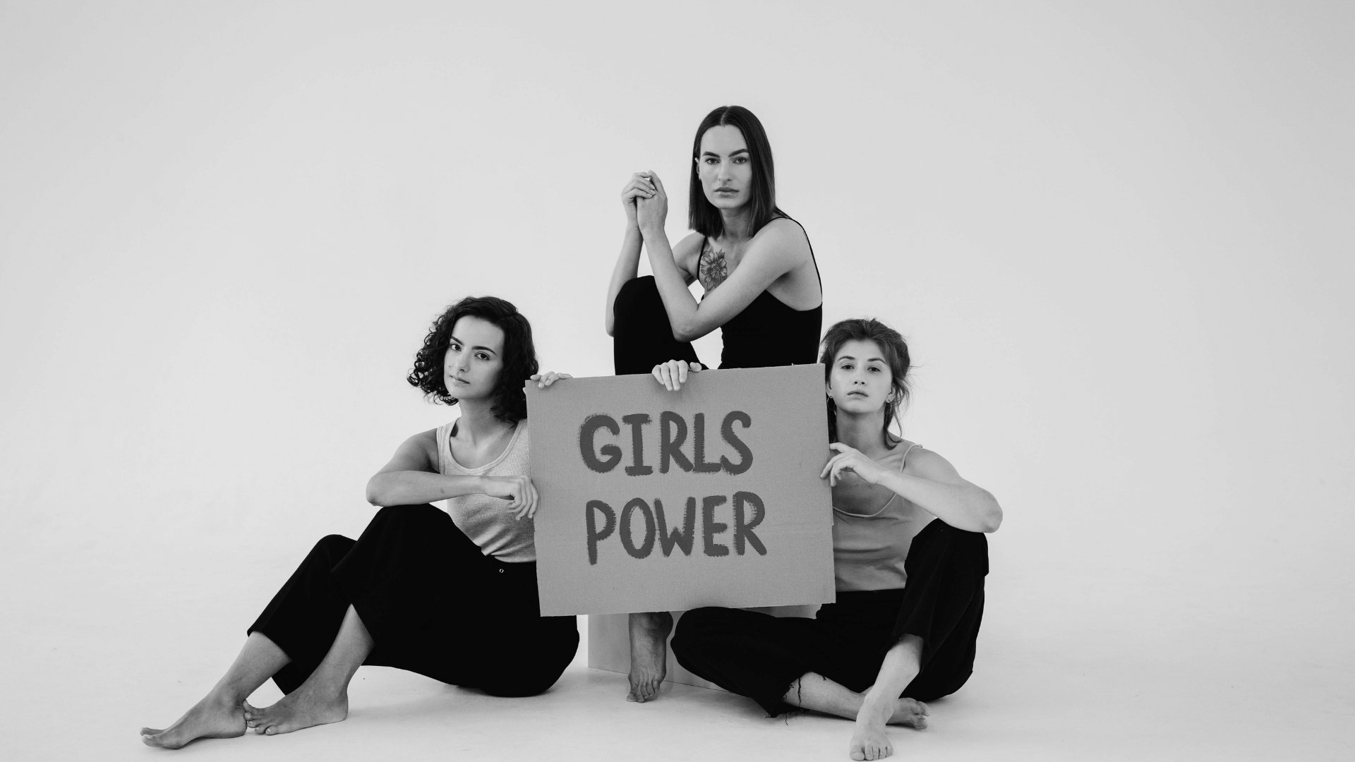 Três mulheres sentadas com as expressões sérias segurando uma placa escrita em tradução literal do inglês 'poder feminino'