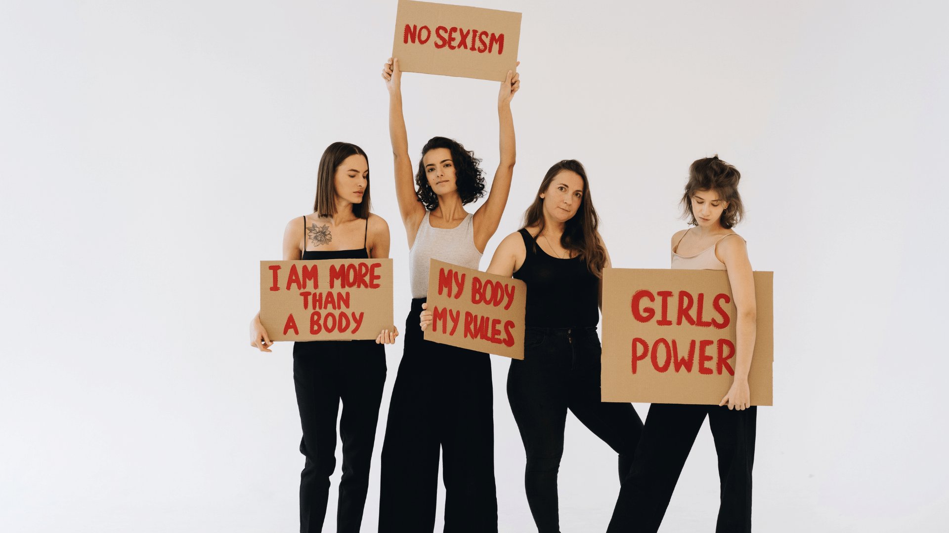 Grupo de quatro mulheres, cada uma segurando um cartaz com os respectivos escritos em tradução literal do inglês 'Eu sou mais do que um corpo', 'não ao machismo', 'meu corpo, minhas regras', 'poder feminino'