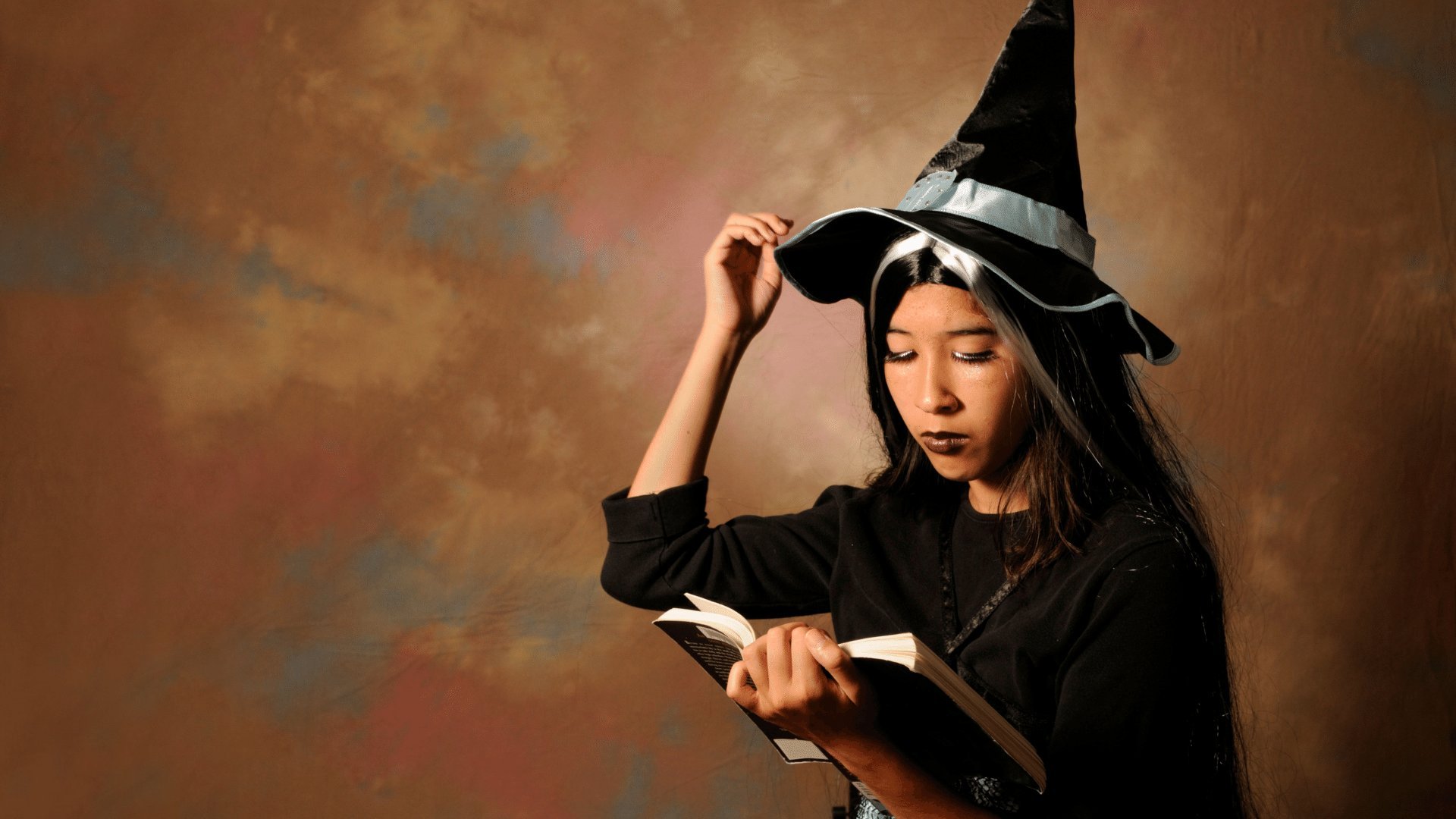 Mulher sentada usando fantasia de bruxa lendo um livro que segura em uma mão e arrumando seu chapéu de bruxa com outra