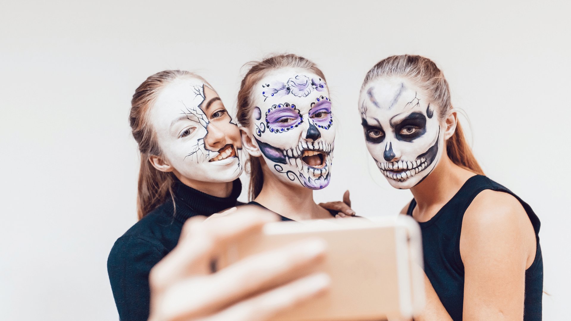 Três garotas com o rosto maquiado de forma a imitar caveiras fazendo caretas para a câmera do celular na qual tiram fotos