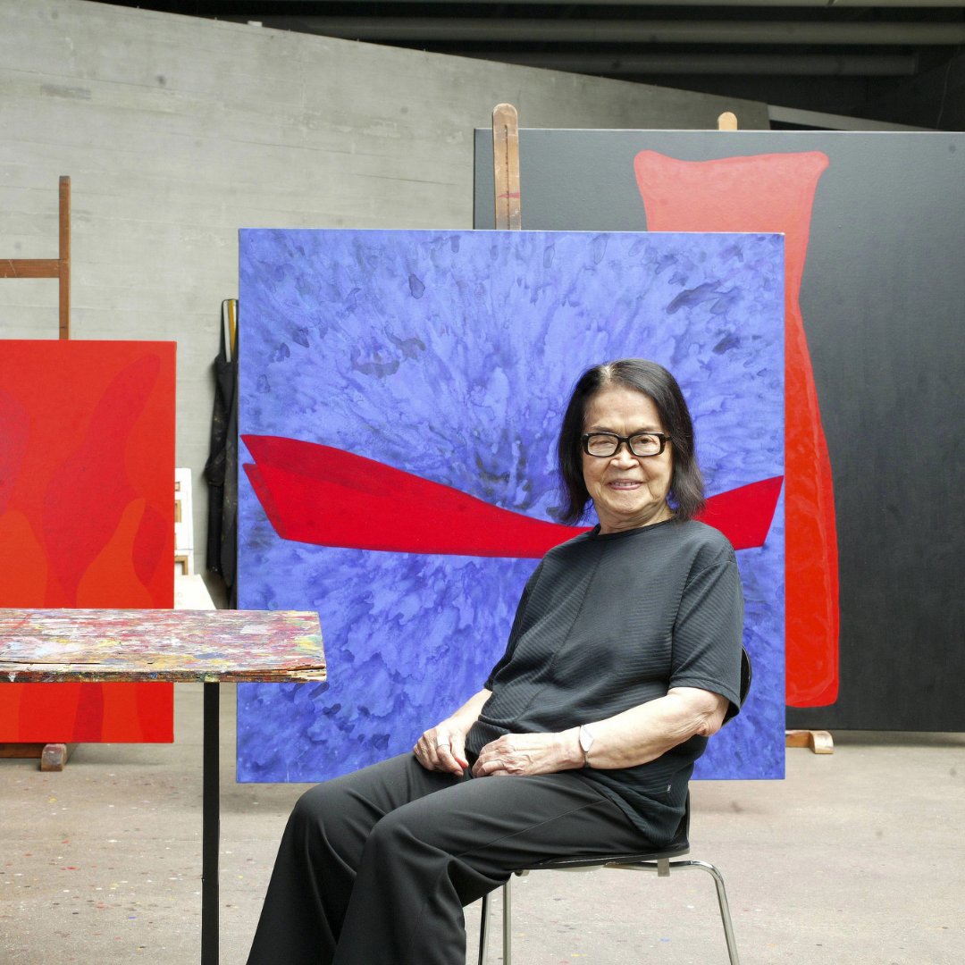Imagem da pintora Tomie Ohtake sentada em frente de alguns quadros