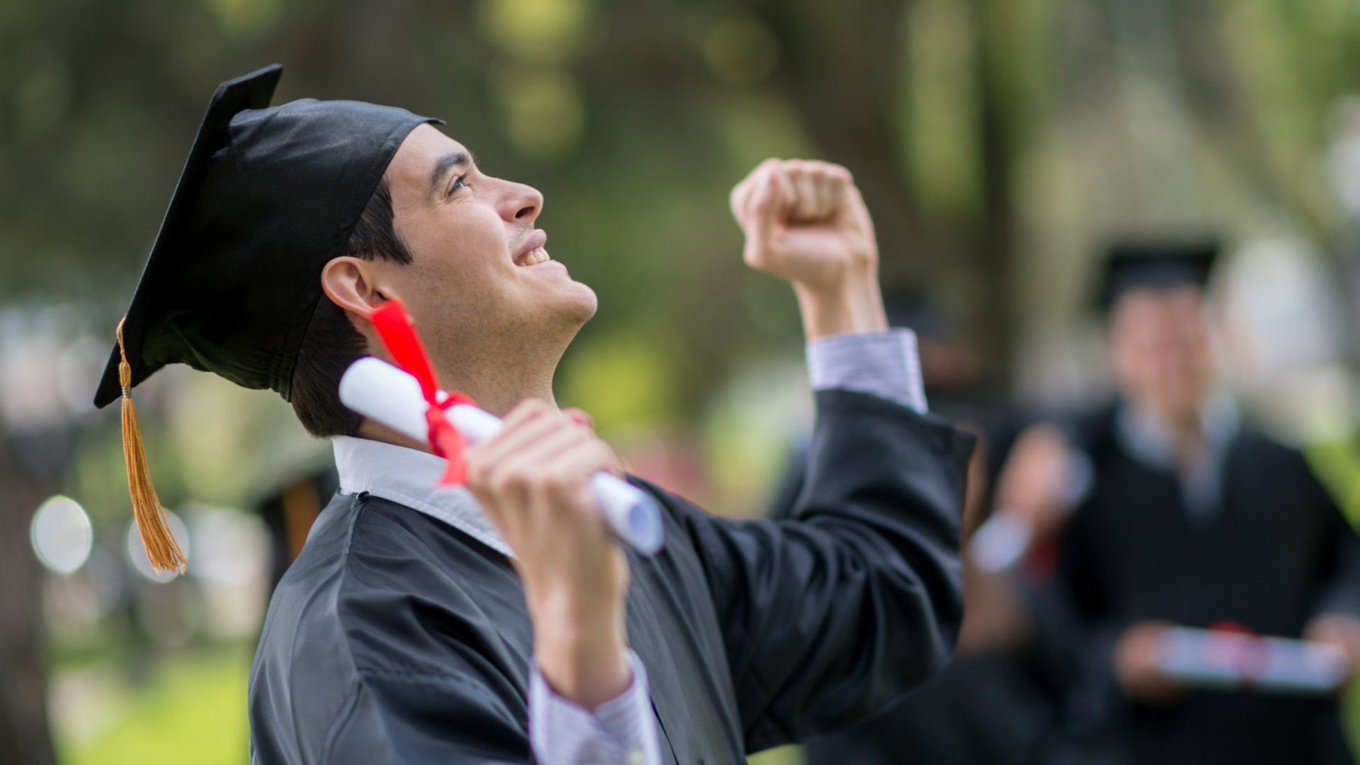 Homem olhando para o alto e sorrindo com os braços erguidos para cima segurando seu diploma
