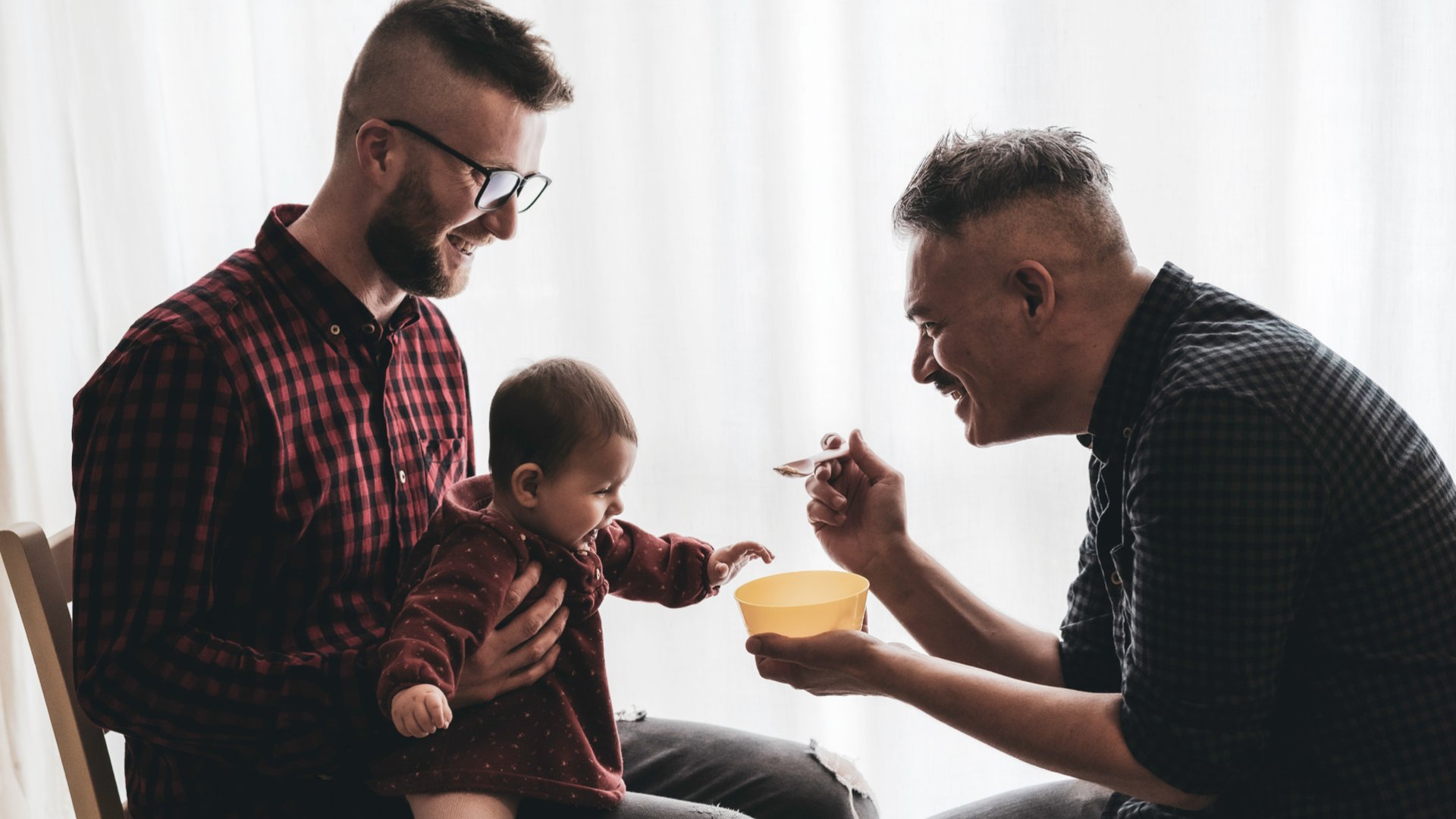 Um homem sentado segurando um neném no colo enquanto outro homem da comida para o bebê