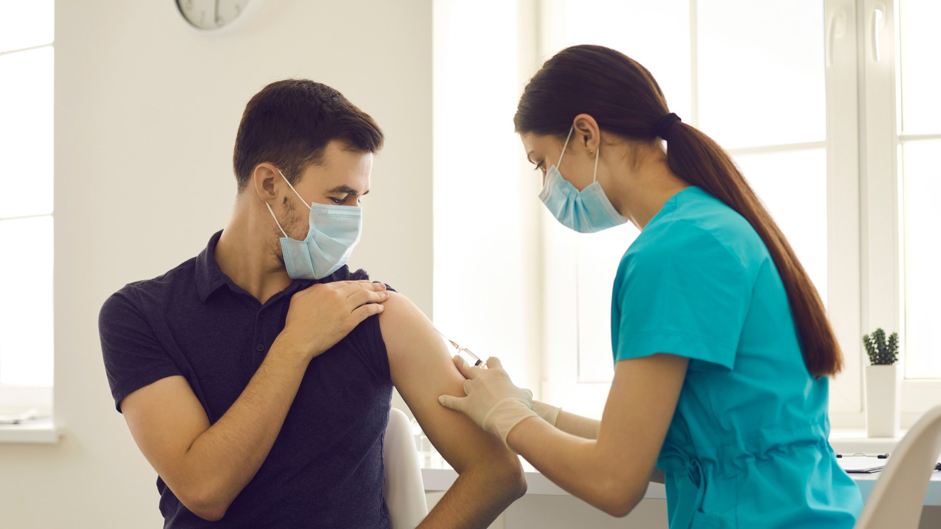 Homem de máscara recendo uma dose de vacina no seu braço esquerdo por uma mulher também de máscara