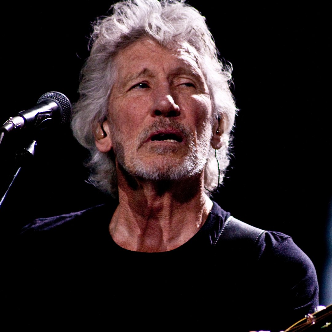 Imagem do cantor Roger Waters cantando e tocando guitarra durante um show