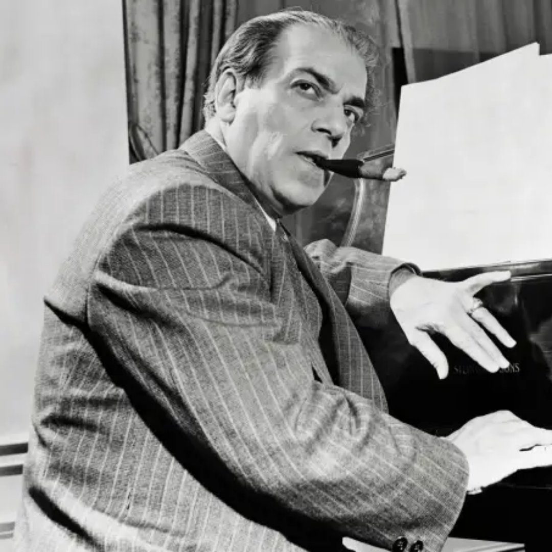 Imagem em preto e branco do maestro Heitor Villa-Lobos fumando um charuto e sentado na frente de um piano