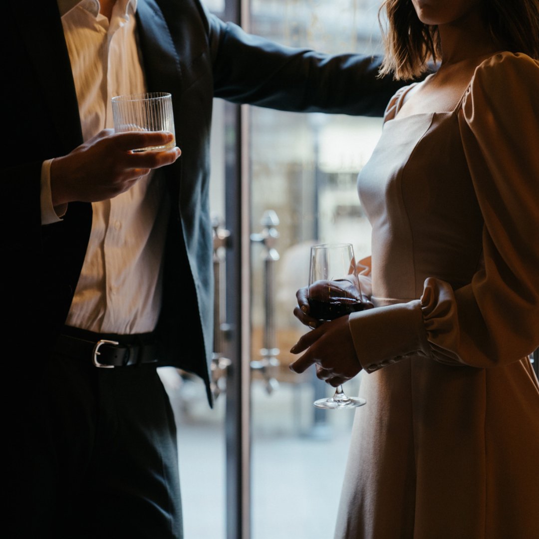 Um homem e uma mulher segurando taças e copos de vinho. Eles conversam.