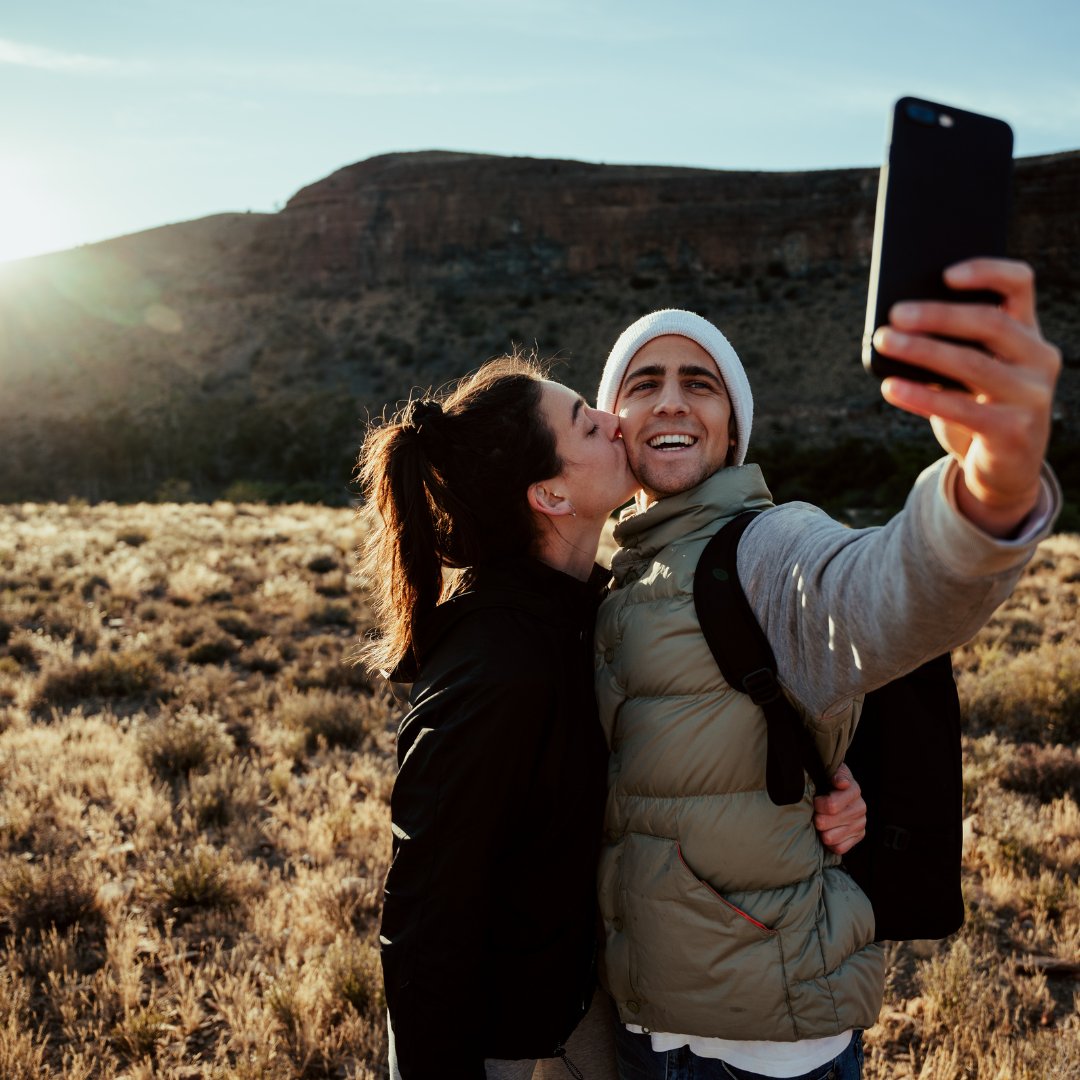 Um homem e uma mulher. A mulher beija a bochecha esquerda do homem e, por sua vez, o homem registra uma selfie. Ambos aparentam estar numa montanha.