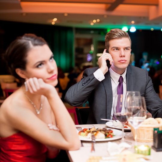 Um homem falando ao telefone durante um encontro com uma mulher. Esta aparenta estar entediada.
