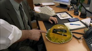 Grampeador do Dwight dentro de uma gelatina amarela, sobre a mesa de trabalho no escritório. Na cana, uma mão masculina aparece apontando para a gelatina.