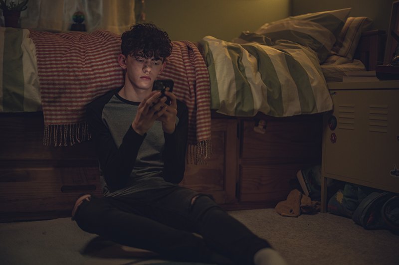 Charlie Spring sentado no chão de seu quarto, recostado na cama e segurando o celular à sua frente.