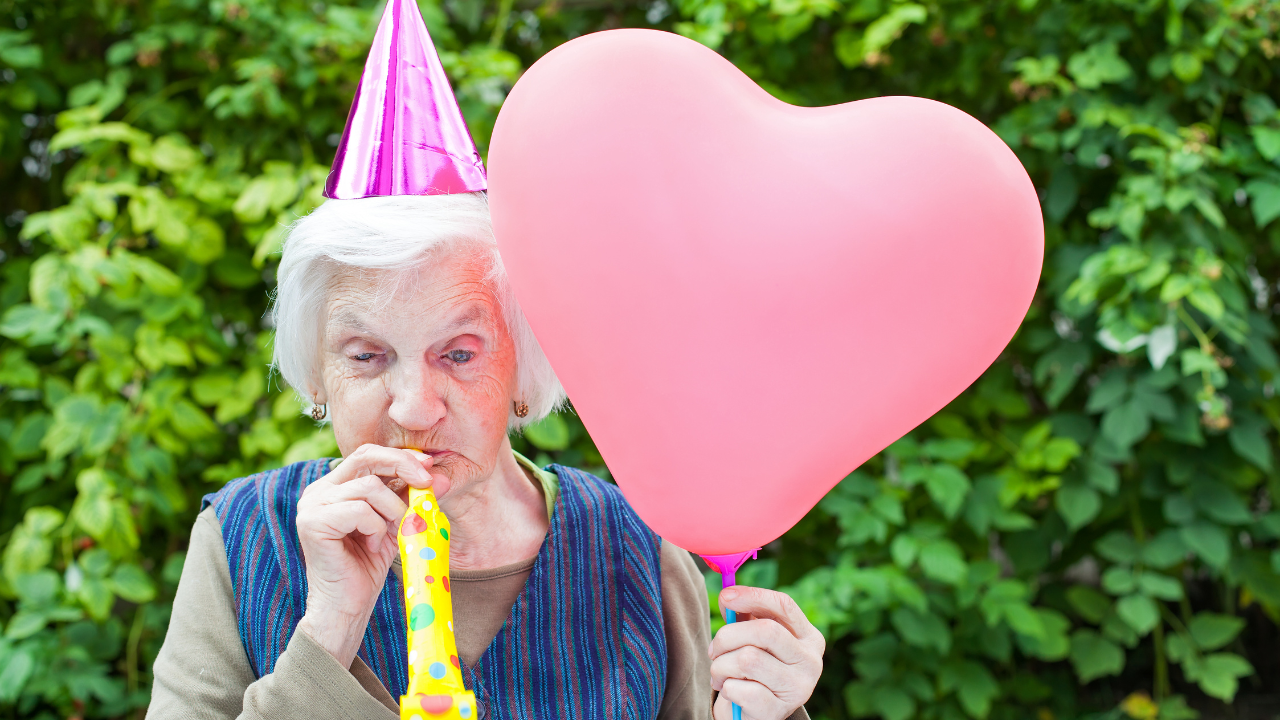 Idosa comemorando o aniversário assoprando uma língua de sogra e segurando um balão em forma de coração
