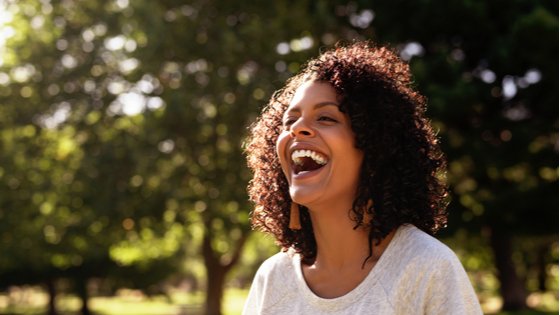 Mulher jovem rindo em um dia ensolarado, com árvores ao fundo.