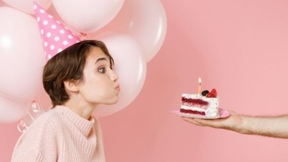 ViDI Studio / Shutterstock Braço segurando prato com uma fatia de bolo e vela acesa e mulher assoprando a vela.