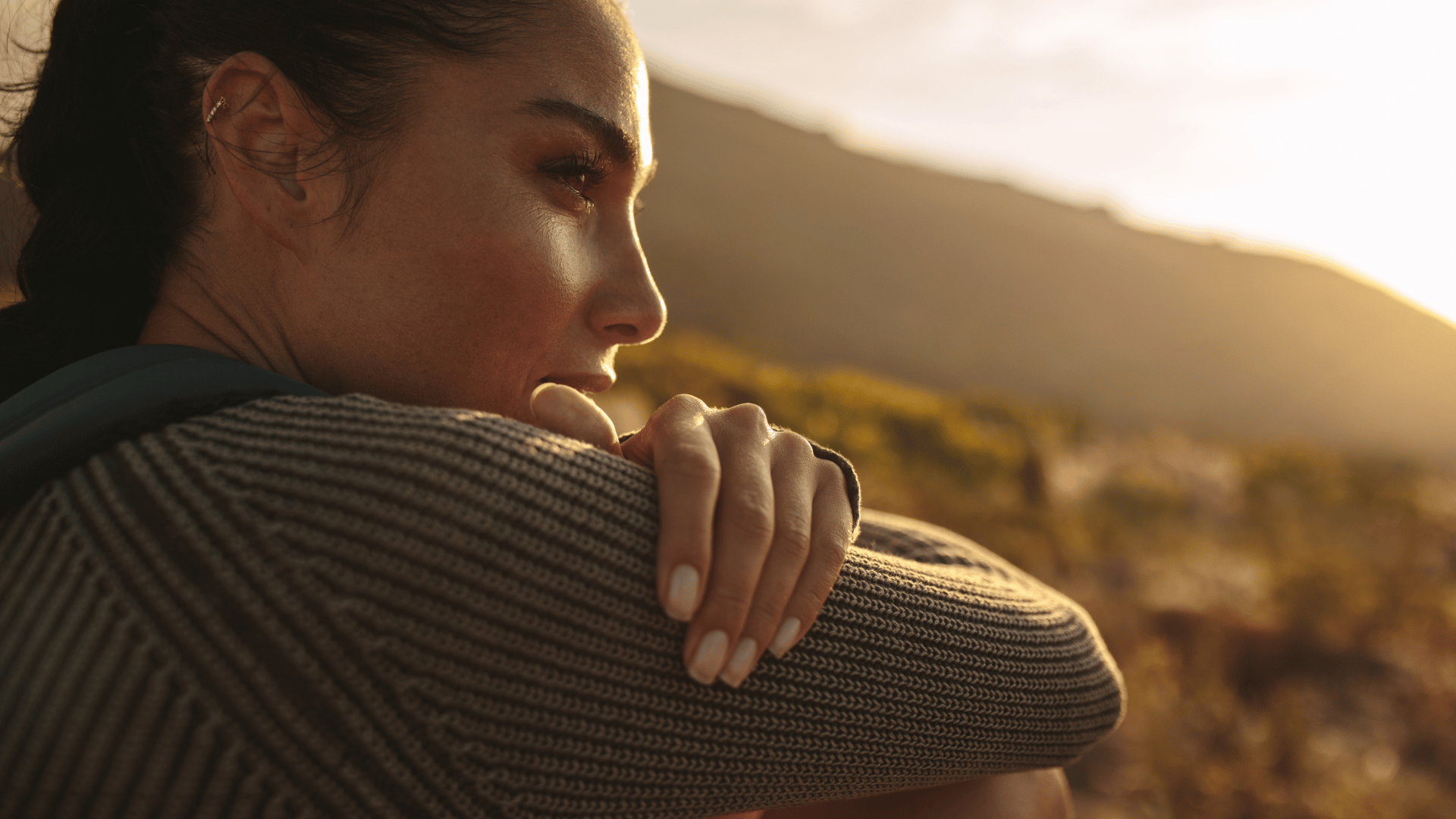 Mulher com uma blusa de tricot marrom, olhando para o infinito. Ao fundo, a imagem de uma montanha e o pôr do Sol.