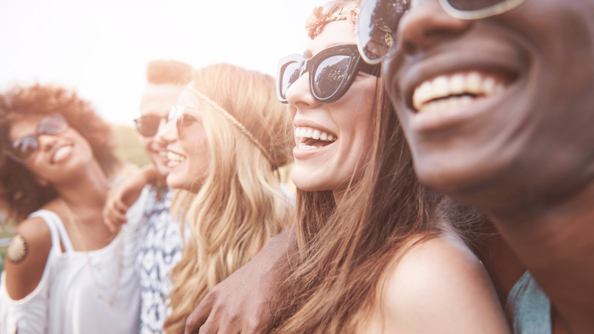 Cinco amigos reunidos, sendo três mulheres e dois homens. Todos estão abraçados, olhando para o infinito. Eles usam óculos de sol e estão felizes e sorridentes.