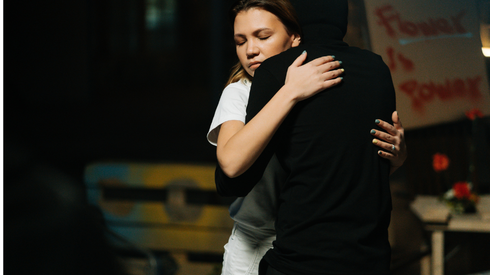 Homem e mulher se abraçando. O homem está de costas e a mulher, de olhos fechados