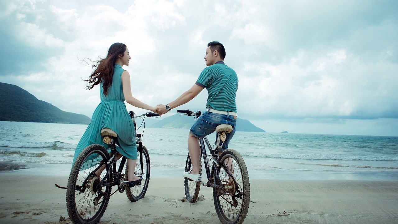 Casal andando de bicicleta pela praia de mãos dadas
