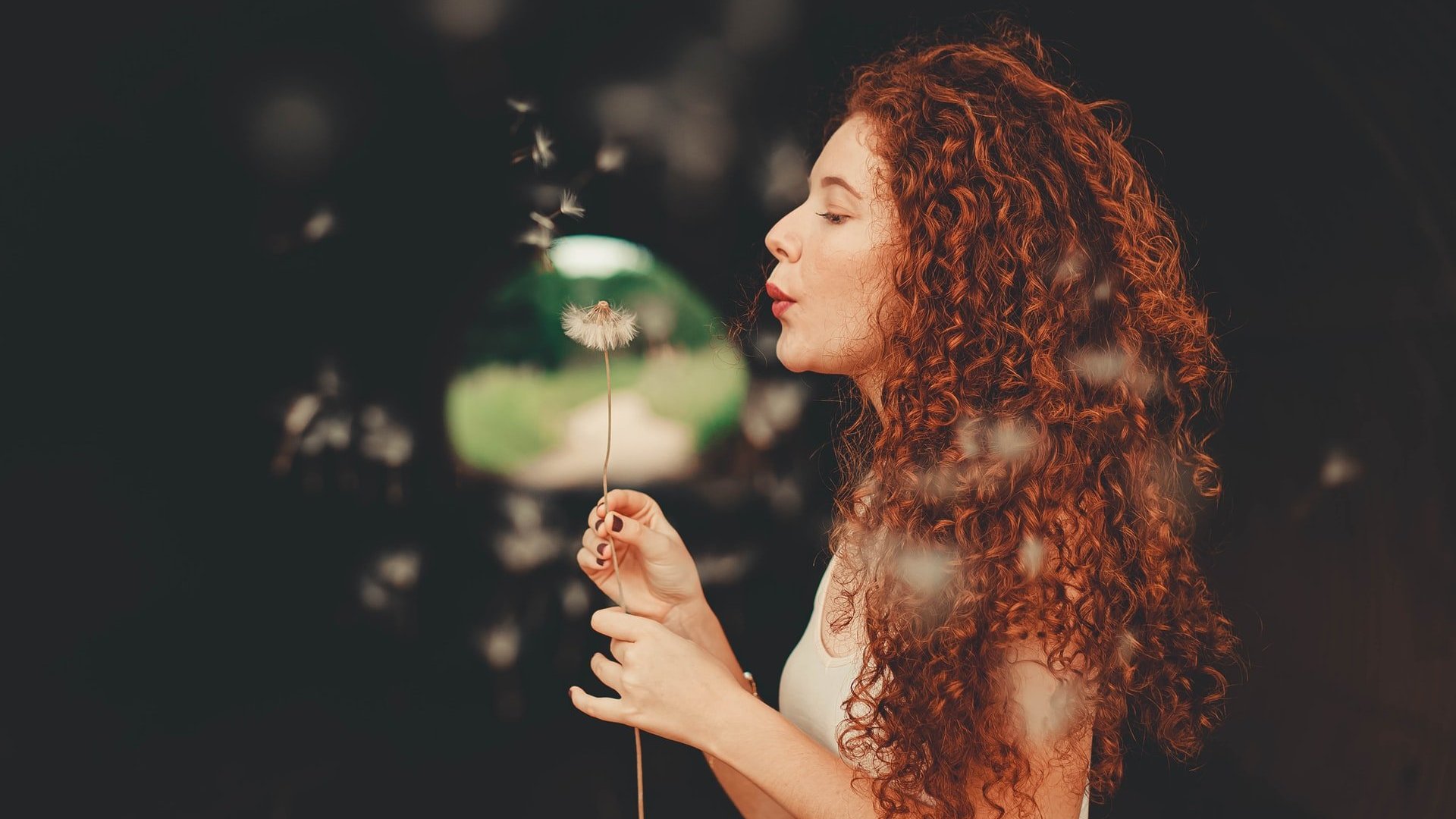 Letícia Pelissari / Unsplash Mulher branca e ruiva assoprando flor dente-de-leão.