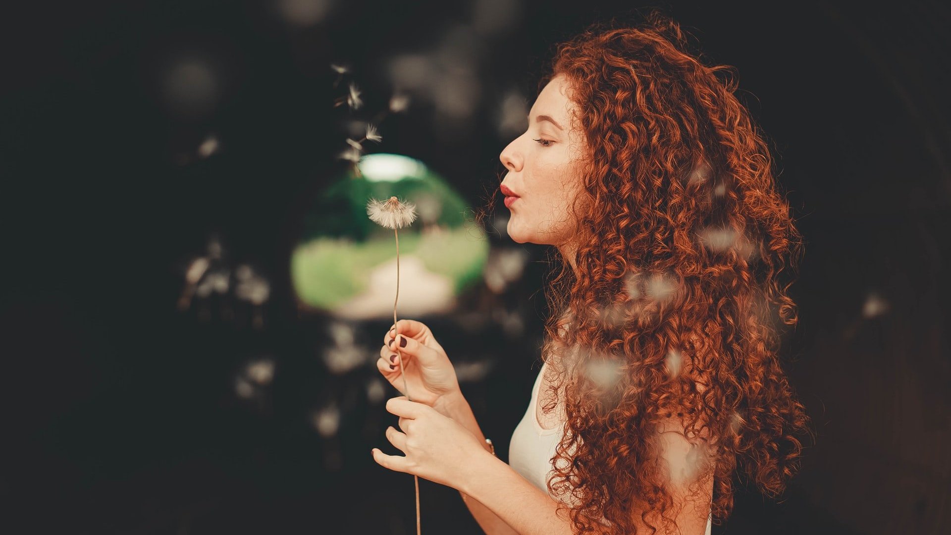 Mulher branca e ruiva assoprando flor dente-de-leão.