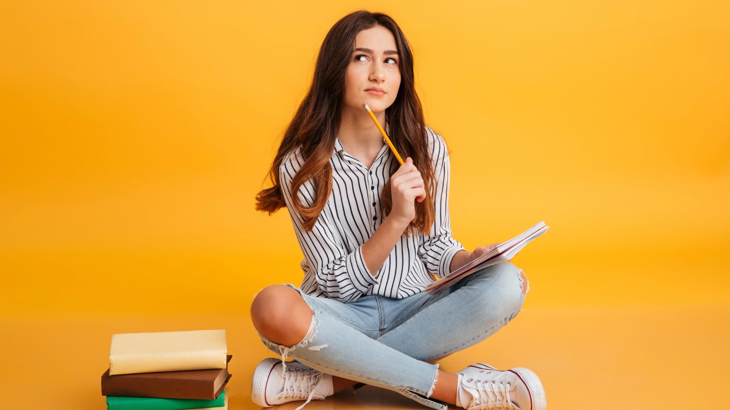 Menina sentada no chão enquanto segura um lápis e um caderno.