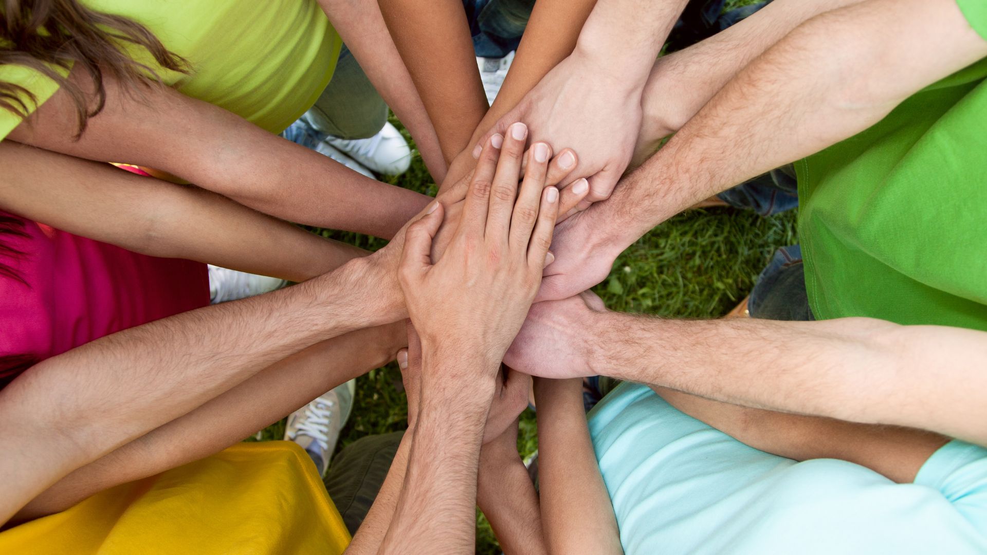 Imagem de vários amigos reunidos com as mãos estendidas, uma sobre a outra. Eles vestem camisetas coloridas.