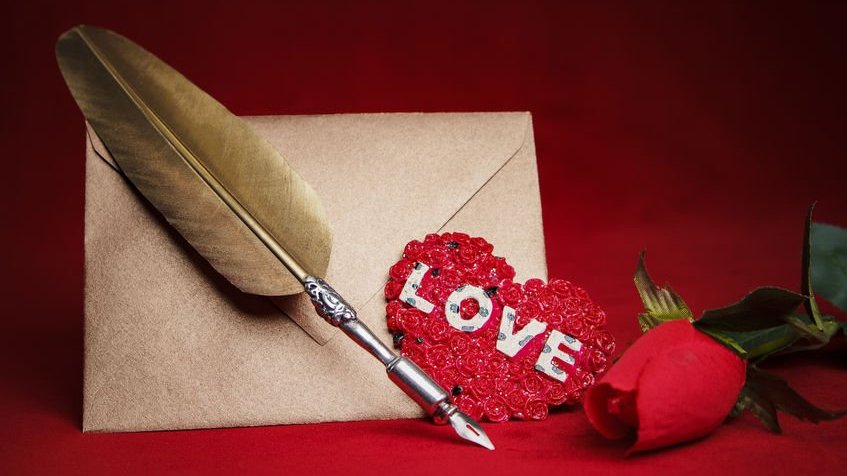 Envelope, pena, coração feito de papel e rosa vermelha, tudo em cima de uma mesa vermelha