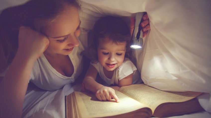 Mãe e filha embaixo de lençol lendo livro de histórias com lanterna