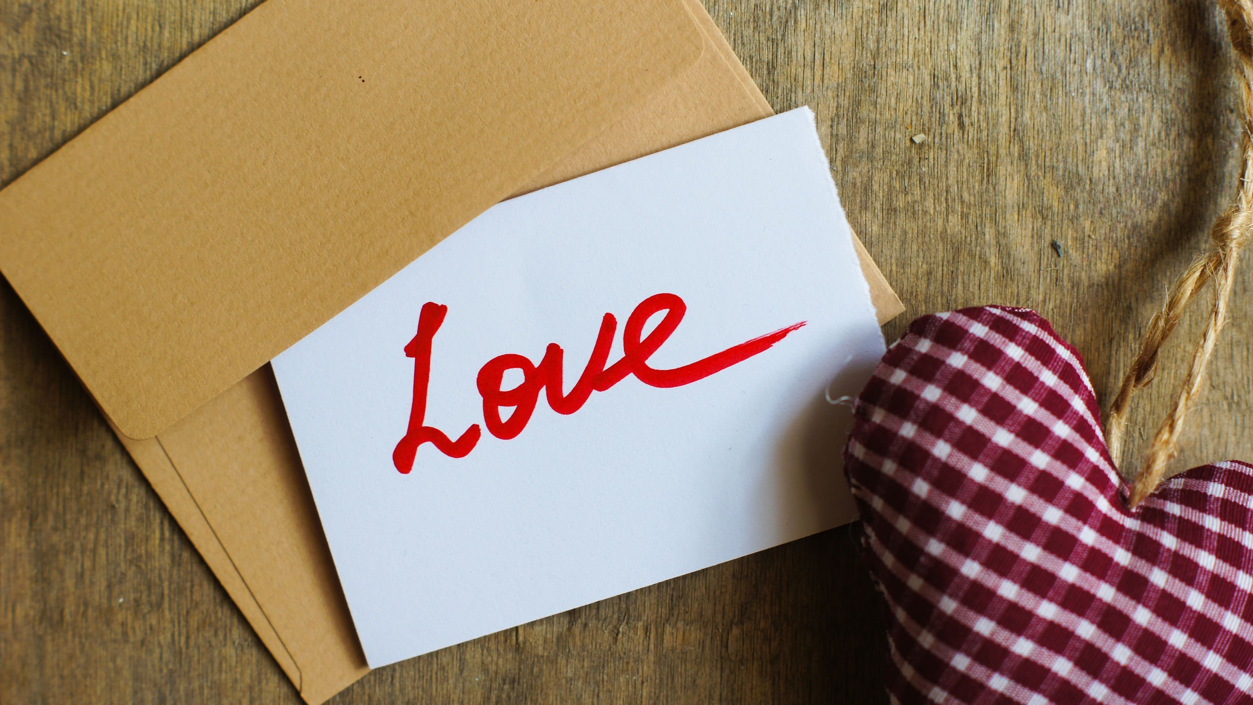 Cartão branco com a palavra amor escrita em vermelho e em inglês, sobre envelope e mesa de madeira, ao lado de coração de pelúcia