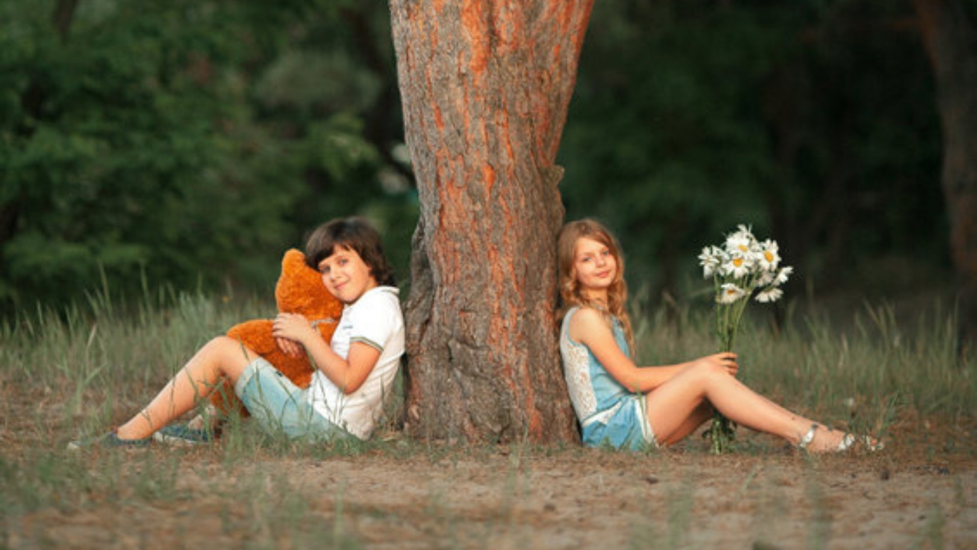 Duas crianças sentadas embaixo de uma àrvore, cada uma de um lado. A menina segura um buquê de flores e o menino um urso de pelúcia