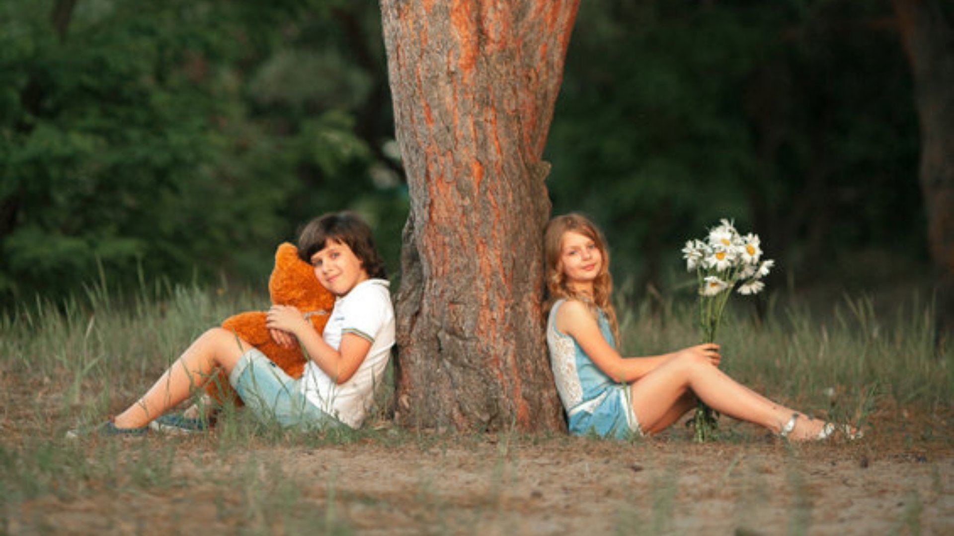 Stasia04 / 123rf Duas crianças sentadas embaixo de uma àrvore, cada uma de um lado. A menina segura um buquê de flores e o menino um urso de pelúcia