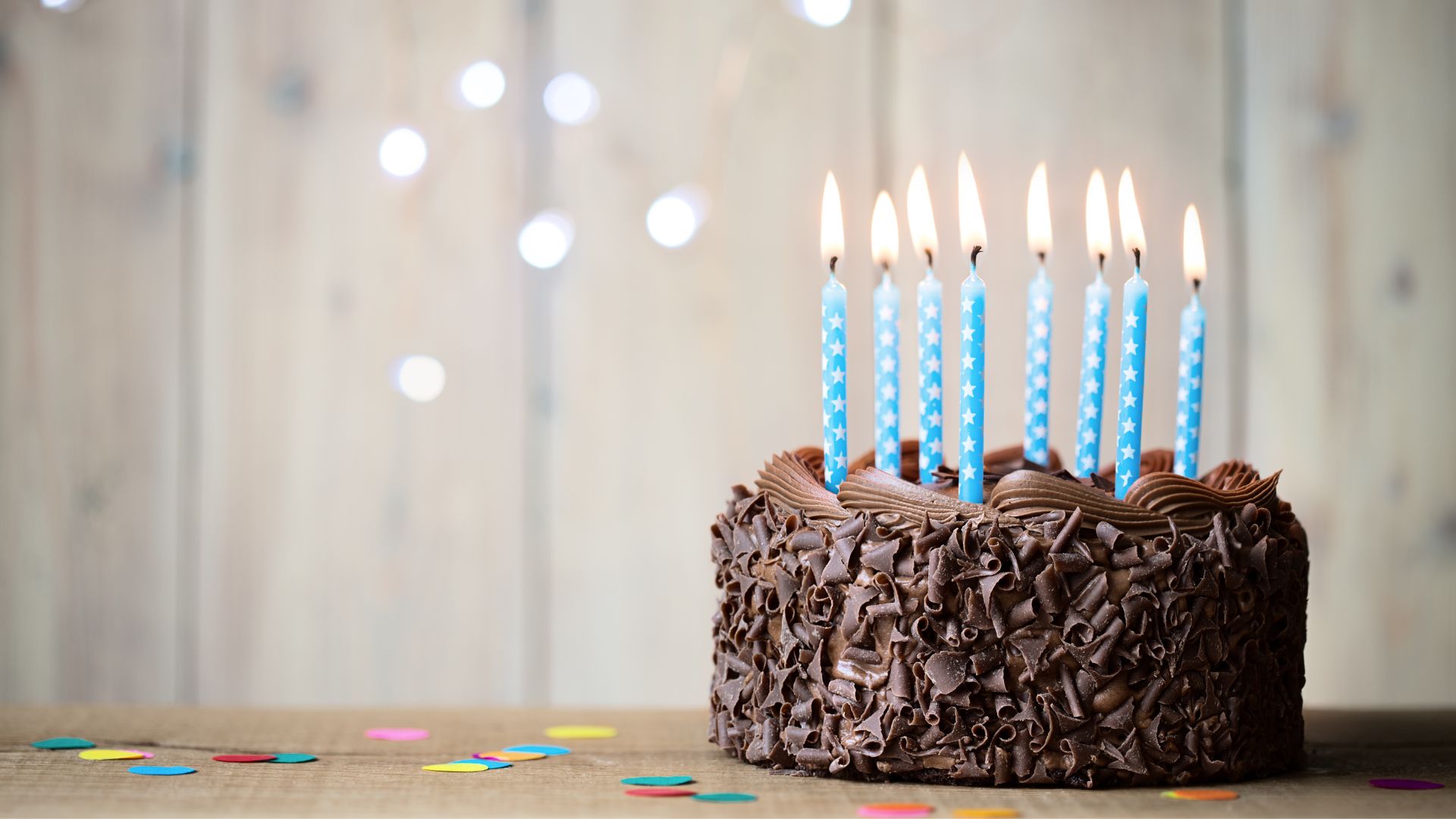 Imagem de um bolo de chocolate com velas azuis no topo, confetes pela mesa e luzes atrás