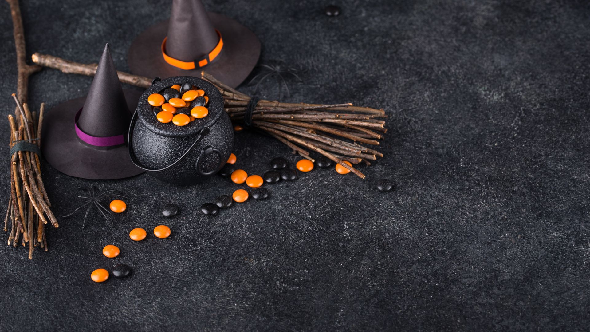 Imagem de fundo preto. Em destaque, acessórios utilizados pelas bruxas como chapéus, caldeirão, gravetos, aranha e pedras nas cores laranja e preto.