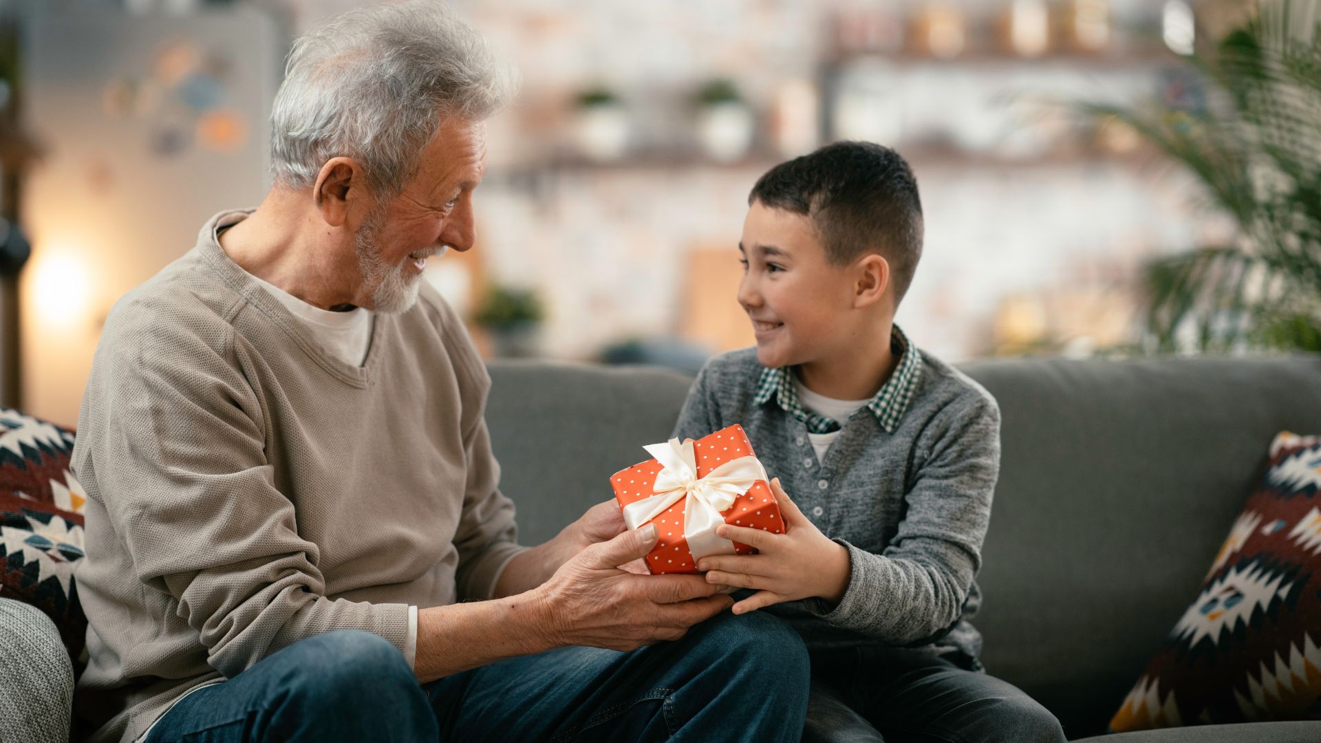 Imagem do avô e do neto sentados em um sofá cinza. O neto está dando uma caixa de presente para o seu avô por conta do seu aniversário.