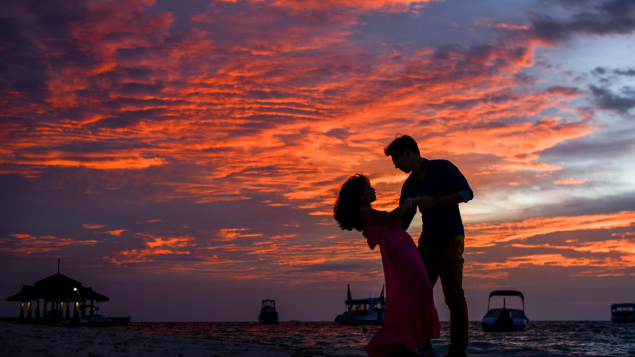 Silhueta de casal dançando. Ao fundo, há um pôr do sol com algumas nuvens na frente