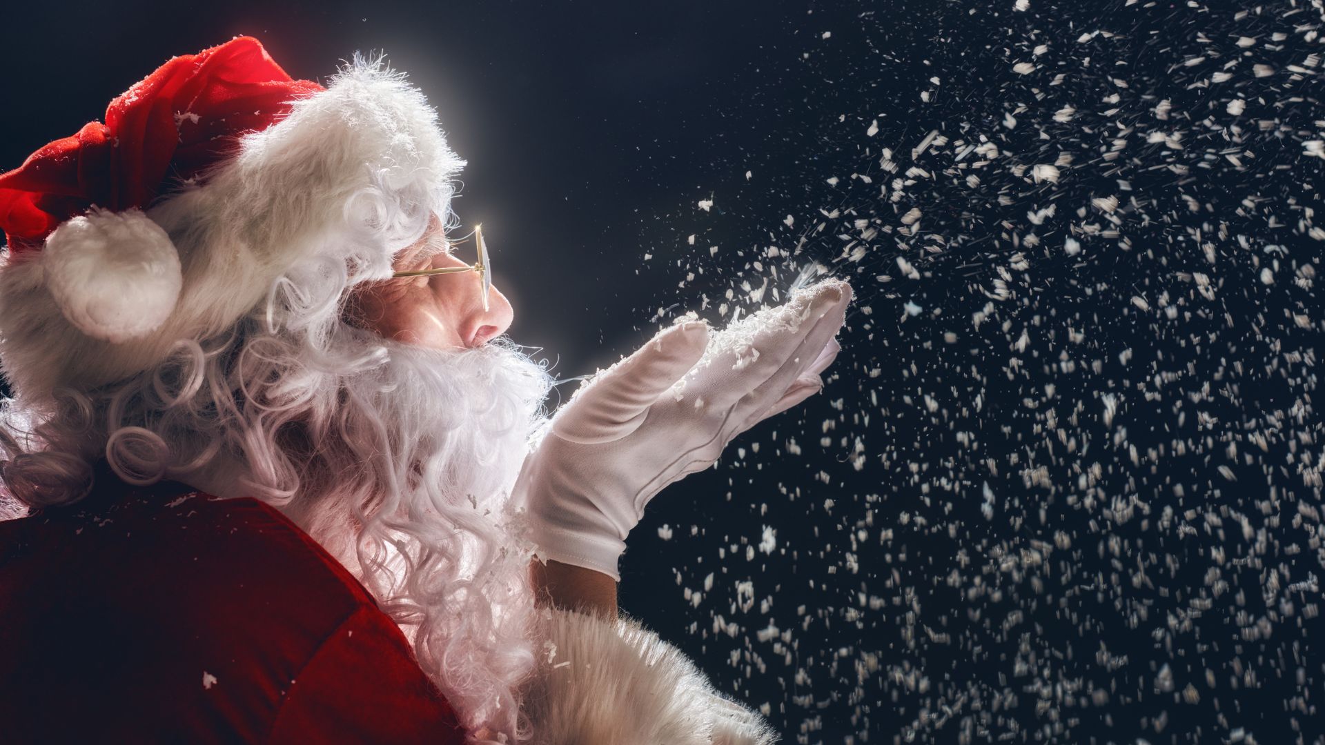 Imagem de um papel noel vestido com sua tradicional roupa e chapéu vermelho assoprando flocos de neve.