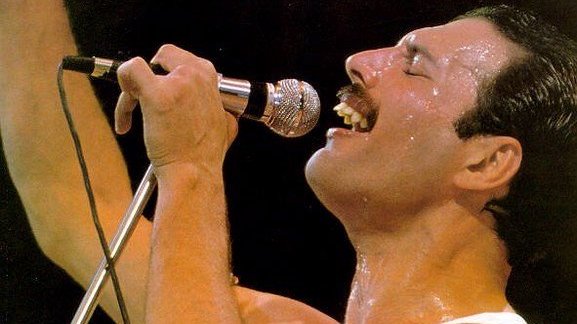 Freddie Mercury segurando seu microfone prateado com olhos fechados e cantando