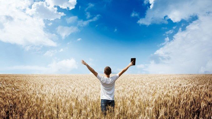Garoto com braços abertos e bíblia em uma das mãos em campo aberto e céu azul