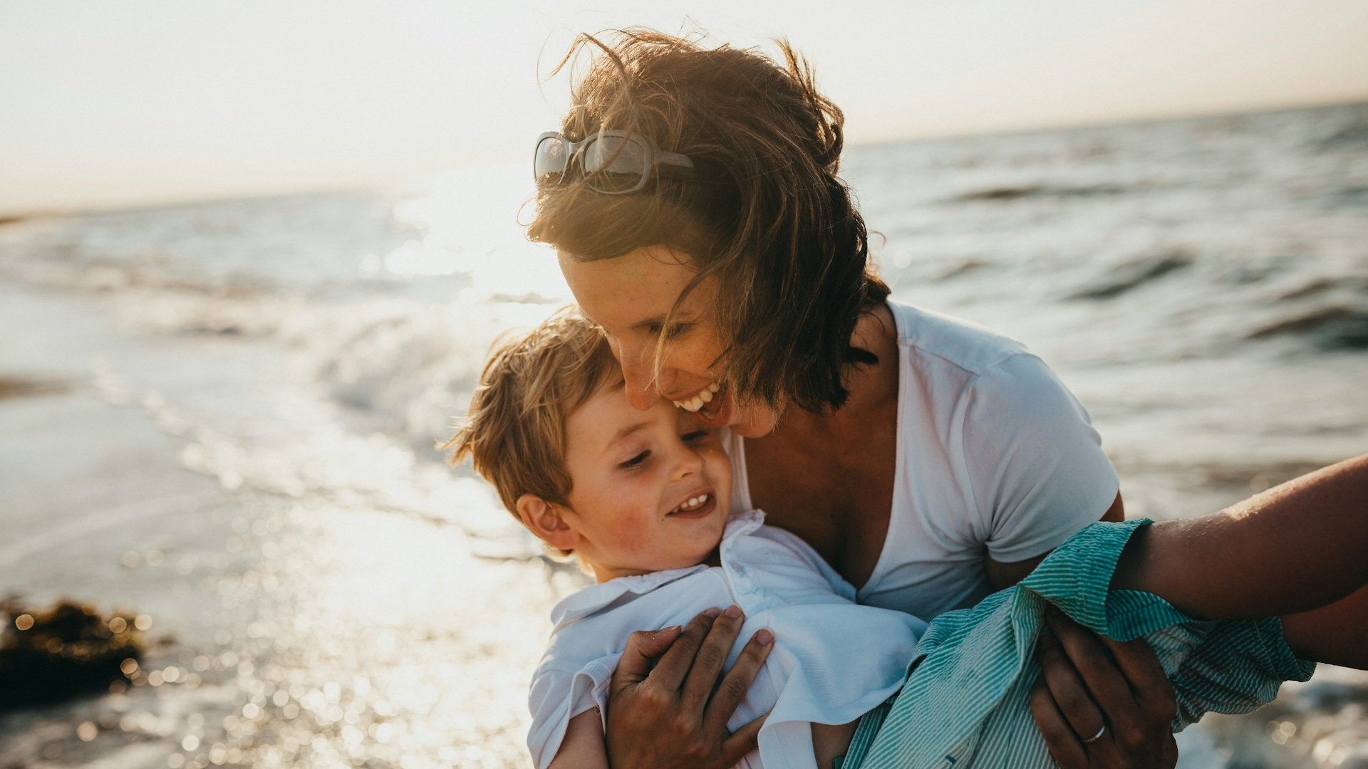 Mãe sorridente com o filho pequeno no colo. Eles estão em uma praia, em frente ao mar.