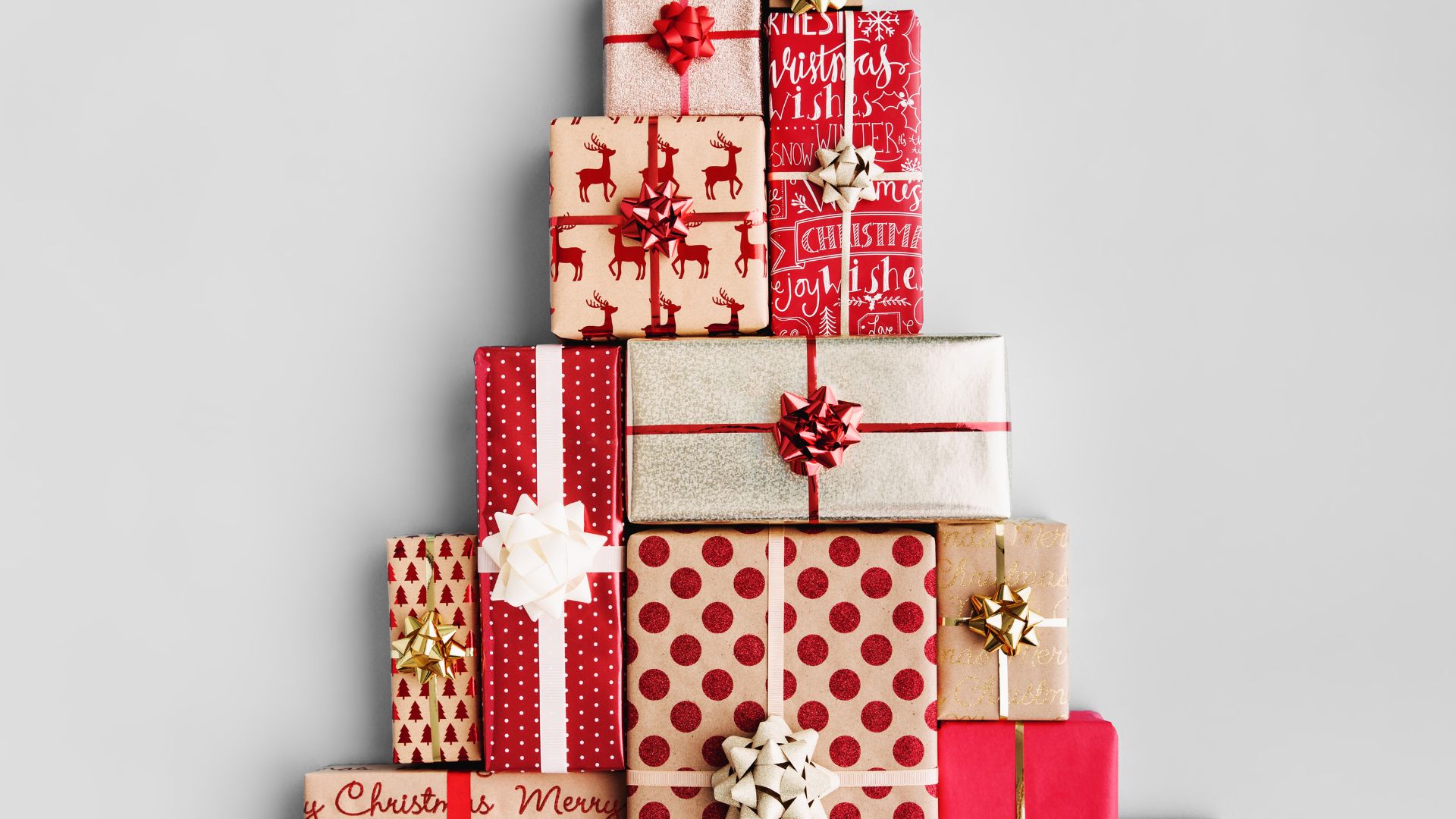 Imagem de fundo branco. Ao centro da mesma, várias caixas de presentes de Natal. Elas estão empilhadas, decoradas com fitas e laços nas cores vermelho, bege e dourado.