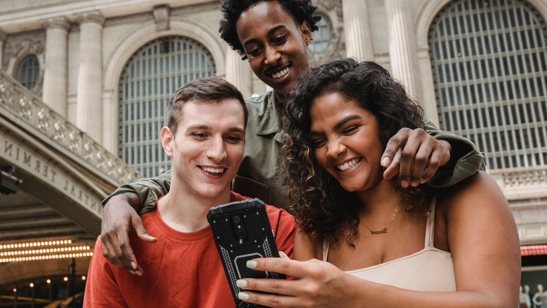 Imagem de três amigos, dois homens e uma mulher, um está de pé abraçando os dois que estão sentados e eles estão sorrindo e olhando no celular