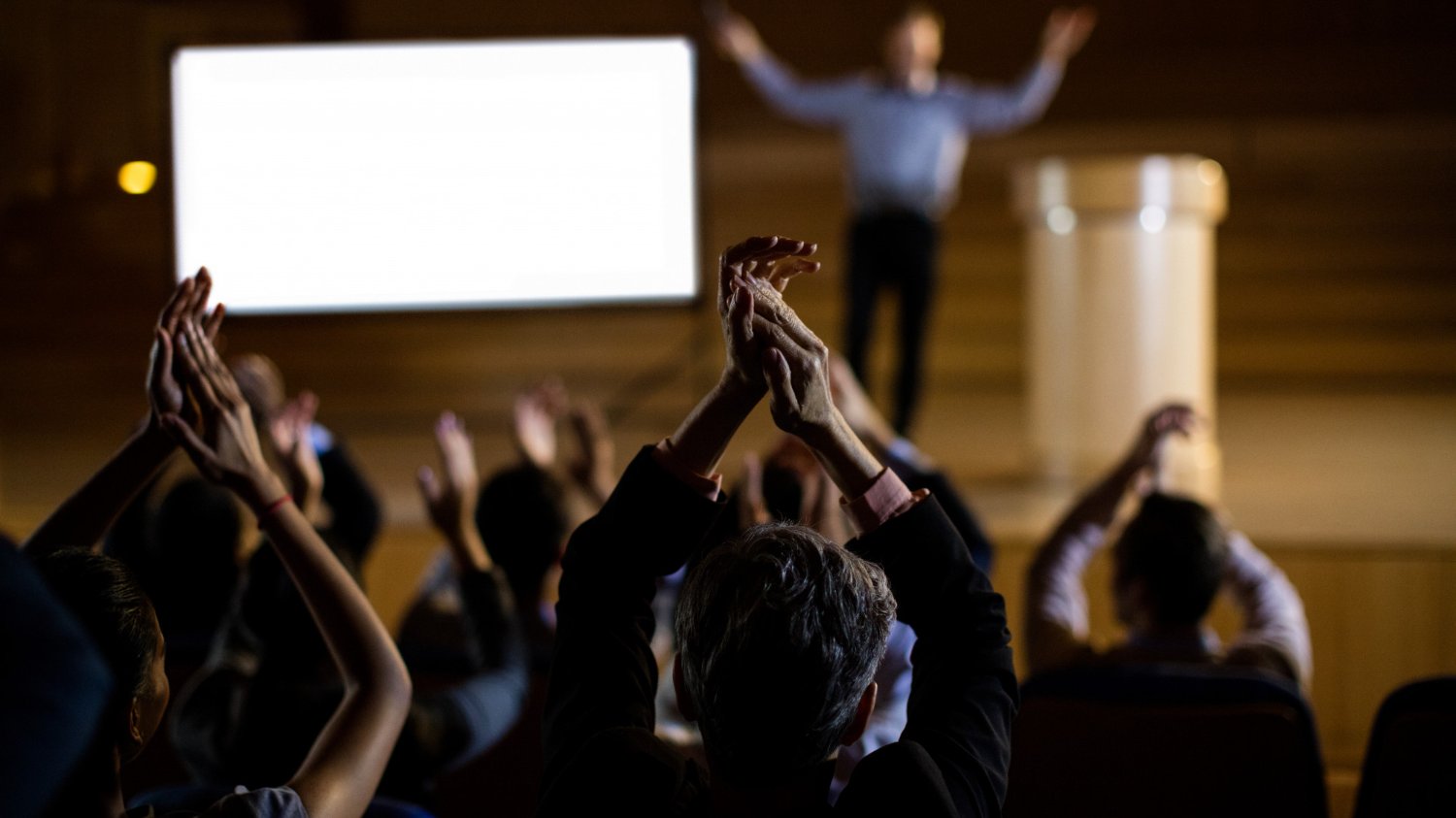 Pessoas aplaudindo uma pessoa em cima de um palco apresentando.