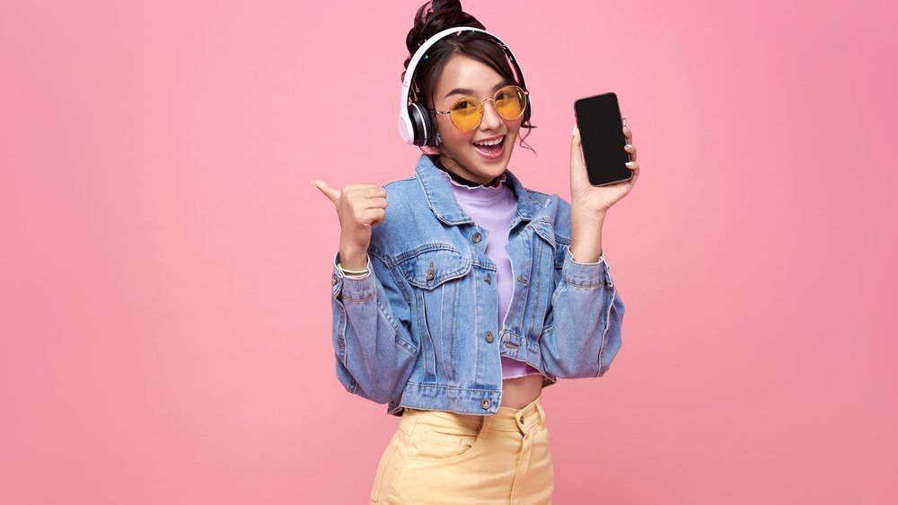 Young Asian teen woman showing smart phone she listening music in headphones isolated on pink background.